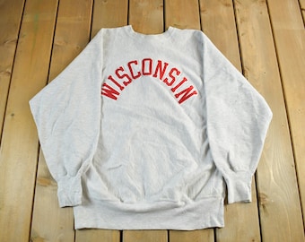 May include: A light gray sweatshirt with the word "WISCONSIN" arched across the chest in red lettering. The sweatshirt has long sleeves and a crew neck. The garment is made of a soft, comfortable material.