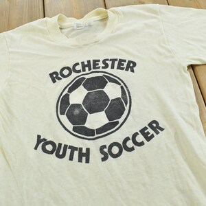May include: A cream-colored short-sleeve t-shirt featuring a black graphic of a soccer ball in the center. The text "ROCHESTER" is printed in a curved arch above the ball, and the text "YOUTH SOCCER" is printed in a curved arch below the ball.
