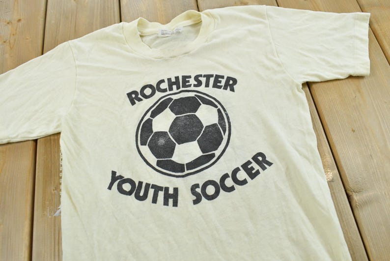 May include: A cream-colored short-sleeve t-shirt featuring a black graphic of a soccer ball in the center. The text "ROCHESTER" is printed in a curved arch above the ball, and the text "YOUTH SOCCER" is printed in a curved arch below the ball.