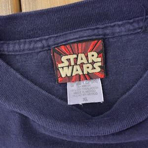May include: A navy blue t-shirt with a red and white Star Wars logo tag. The tag has the text "Star Wars" in white letters. The tag also has the text "100% Cotton", "Made in Mexico", "Hecho en Mexico", and "XL".