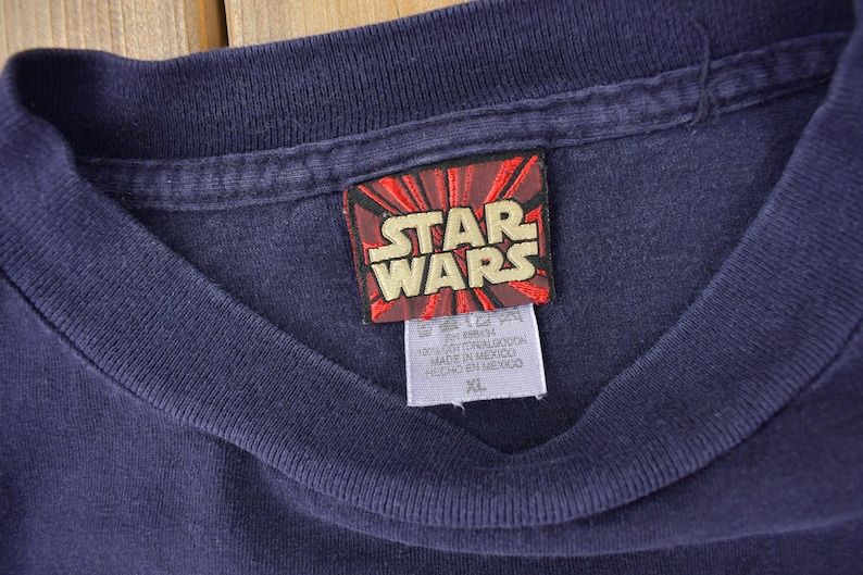 May include: A navy blue t-shirt with a red and white Star Wars logo tag. The tag has the text "Star Wars" in white letters. The tag also has the text "100% Cotton", "Made in Mexico", "Hecho en Mexico", and "XL".