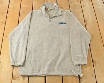 May include: A light gray fleece pullover with a quarter-zip collar and a small blue and teal logo on the chest. The long-sleeved top has a drawstring at the hem and is laid out on a wooden surface.