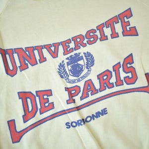 May include: A yellow long-sleeve shirt with the text "UNIVERSITE DE PARIS" in red and blue lettering. The text is in a curved arc with a crest in the center. The text "SORBONNE" is printed in blue below the arc.