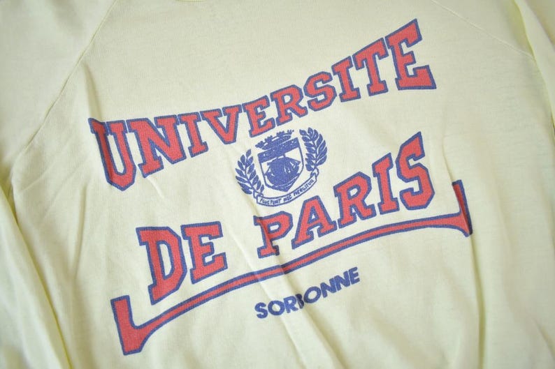 May include: A yellow long-sleeve shirt with the text "UNIVERSITE DE PARIS" in red and blue lettering. The text is in a curved arc with a crest in the center. The text "SORBONNE" is printed in blue below the arc.
