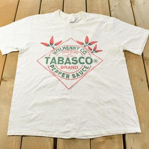 以下が含まれることがあります： タバスコブランドのロゴが入った白いTシャツ。ロゴはひし形で、緑と赤で「TABASCO BRAND PEPPER SAUCE」と書かれています。シャツはクルーネックで半袖です。