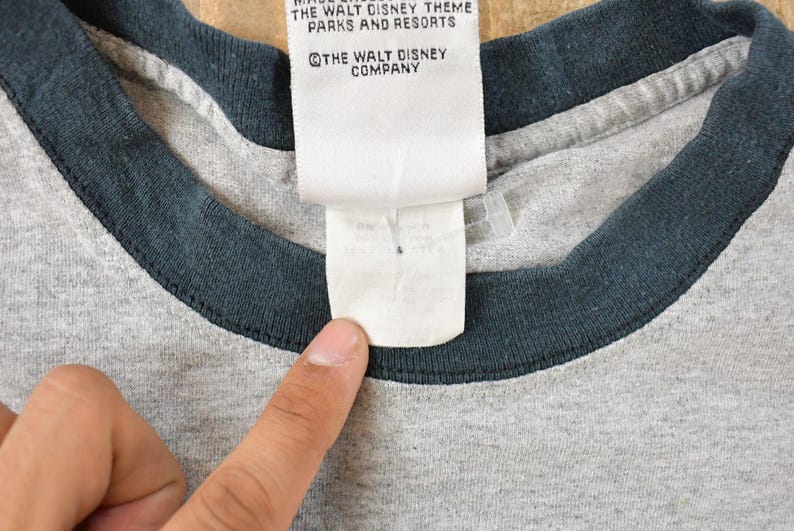 May include: Close-up of a gray t-shirt with a dark blue collar. A white tag with text from The Walt Disney Company is visible. The shirt is made of a soft, comfortable material.