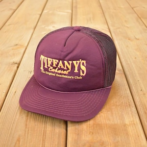Op de afbeelding: Bordeaux truckerpet met een mesh achterkant en een gebogen klep. De voorkant van de pet toont de tekst "TIFFANY'S Cabaret The Original Gentlemen's Club" in gouden letters. De pet ligt op een houten oppervlak.