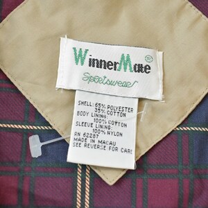 May include: Close-up of a jacket label with the brand "Winner Mate Sportswear". The label is white with green text and is sewn onto a tan fabric patch. The jacket lining is a plaid pattern in burgundy, navy, and green.