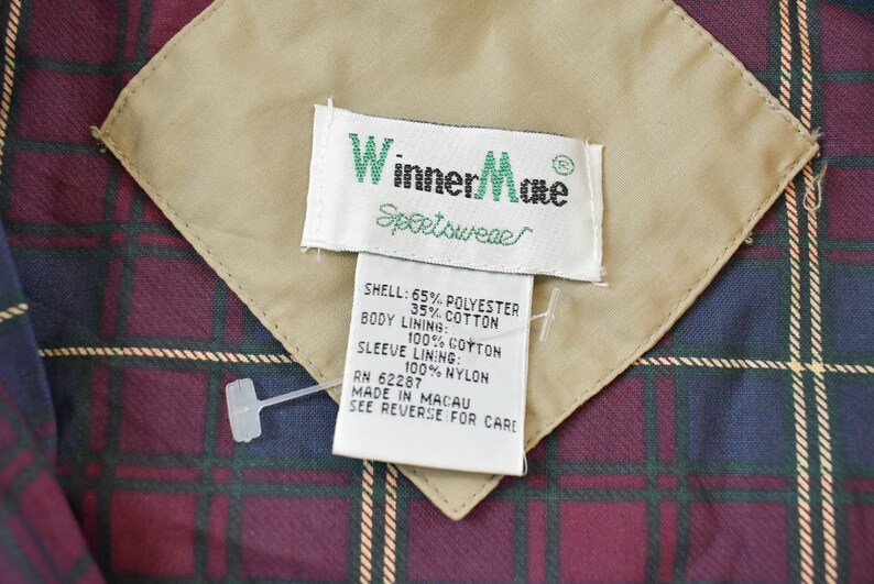 May include: Close-up of a jacket label with the brand "Winner Mate Sportswear". The label is white with green text and is sewn onto a tan fabric patch. The jacket lining is a plaid pattern in burgundy, navy, and green.