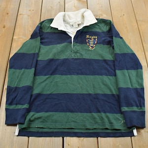 May include: A long-sleeve rugby shirt featuring horizontal navy blue and dark green stripes. The shirt has a white collar, a button-up placket, and an embroidered crest on the chest with the word Rugby and three small yellow crowns.