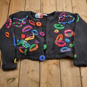 May include: A black cardigan sweater with colorful embroidered circles and lines. The cardigan has a button closure.