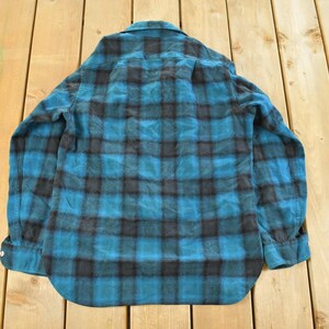May include: A teal and black plaid flannel shirt with long sleeves. The shirt is buttoned and has a collar. The shirt is lying on a wooden surface.