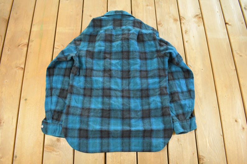 May include: A teal and black plaid flannel shirt with long sleeves. The shirt is buttoned and has a collar. The shirt is lying on a wooden surface.