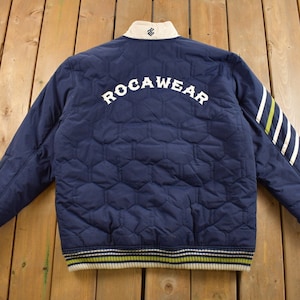 Puede incluir: Una chaqueta acolchada azul marino con un logotipo "ROCAWEAR" blanco en la espalda. La chaqueta tiene rayas blancas y verdes en las mangas y una banda rayada blanca y verde alrededor de la parte inferior.