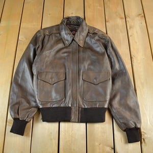 Puede incluir: Chaqueta bomber de cuero marrón con cuello, dos bolsillos delanteros y puños y dobladillo de canalé negros. La chaqueta se exhibe sobre una superficie de madera.