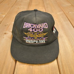 Może przedstawiać: Czarna czapka bejsbolówka z haftowanym logo w kolorze fioletowym i złotym, na którym widnieje napis "Brickyard 400" i "Inaugural Race August 6, 1994".