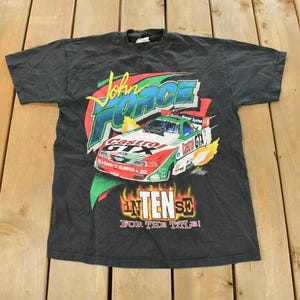 Op de afbeelding: Zwart T-shirt met een raceauto-afbeelding, de tekst "John Force Castrol GTX" en "INTENSE FOR THE TILE!". Het shirt heeft een ronde hals en korte mouwen. De afbeelding is rood, groen en wit.
