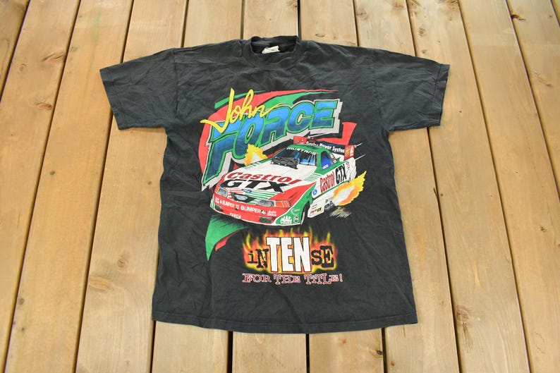 Op de afbeelding: Zwart T-shirt met een raceauto-afbeelding, de tekst "John Force Castrol GTX" en "INTENSE FOR THE TILE!". Het shirt heeft een ronde hals en korte mouwen. De afbeelding is rood, groen en wit.