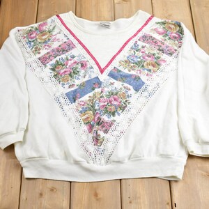 Puede incluir: Sudadera blanca con un diseño floral y de encaje en la parte delantera. El diseño presenta un patrón floral rosa, azul y blanco con un ribete de encaje blanco.
