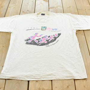 May include: A white short-sleeve t-shirt featuring a 1991 graphic print that reads "MS WALK for Multiple Sclerosis" and "walk in their shoes" with a stylized illustration of multiple figures in pink, blue, and black colors across the chest.