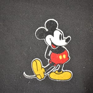 May include: Black hooded sweatshirt featuring a colorful Mickey Mouse graphic. The cartoon character is depicted in his classic pose, with a white face, black ears, red shorts, and yellow shoes. The design is outlined in white.