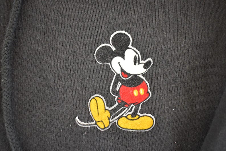 May include: Black hooded sweatshirt featuring a colorful Mickey Mouse graphic. The cartoon character is depicted in his classic pose, with a white face, black ears, red shorts, and yellow shoes. The design is outlined in white.