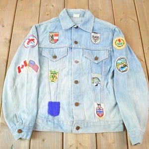May include: A light blue denim jacket with multiple patches sewn on. The patches feature various designs, including flags, logos, and illustrations.