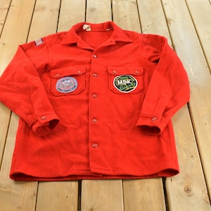 May include: A bright red long-sleeve button-down shirt featuring a pointed collar, two chest pockets with flaps, and buttoned cuffs. The shirt displays a small American flag patch on the right shoulder and two circular embroidered patches on the chest.