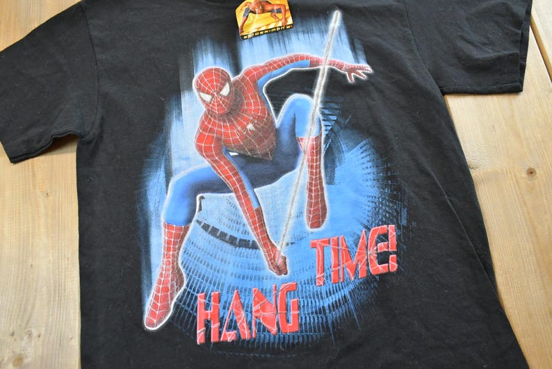 May include: Black t-shirt with a Spider-Man graphic in a dynamic pose, showcasing red and blue costume details. The words "HANG TIME!" are printed in red. A tag is attached at the top.