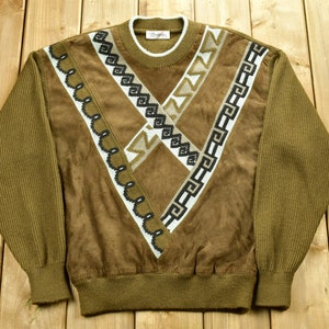 May include: A brown sweater with a geometric pattern in black, white, and beige. The sweater has a suede-like texture and a ribbed collar, cuffs, and hem.