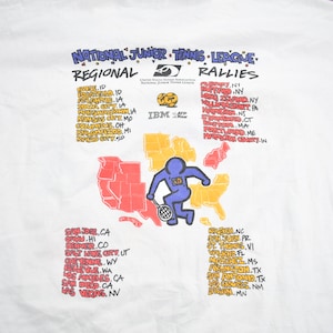 Puede incluir: Una camiseta blanca con un gr&aacute;fico de un jugador de tenis de dibujos animados y un mapa de los Estados Unidos. El texto "National Junior Tennis League Regional Rallies" est&aacute; en la parte superior del gr&aacute;fico. El texto "IBM" est&aacute; en el centro del gr&aacute;fico. El texto enumera ciudades y estados en los Estados Unidos.