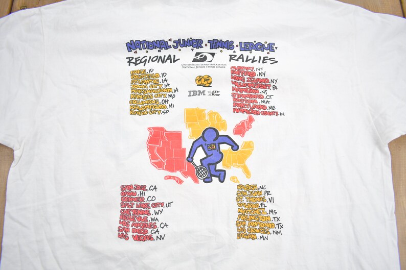 Puede incluir: Una camiseta blanca con un gr&aacute;fico de un jugador de tenis de dibujos animados y un mapa de los Estados Unidos. El texto "National Junior Tennis League Regional Rallies" est&aacute; en la parte superior del gr&aacute;fico. El texto "IBM" est&aacute; en el centro del gr&aacute;fico. El texto enumera ciudades y estados en los Estados Unidos.
