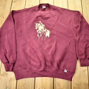 May include: A maroon sweatshirt with a gold embroidered image of a person riding a horse. The sweatshirt has a crew neck and long sleeves.