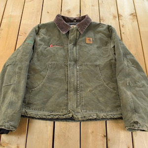 Peut inclure: Veste de travail vert olive avec un col en velours côtelé marron. Comprend deux grandes poches avant, une petite poche poitrine et un écusson Carhartt. La veste a un aspect vintage usé.