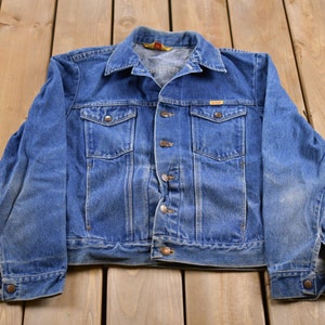 May include: A blue denim jacket with a button closure, two chest pockets, and long sleeves.