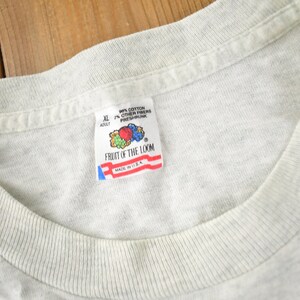 May include: A grey Fruit of the Loom t-shirt tag with the text "90% Cotton 10% Other Fibres Preshrunk" and "Fruit of the Loom" with a logo of fruit. The tag also says "XL Adult" and "Made in U.S.A."