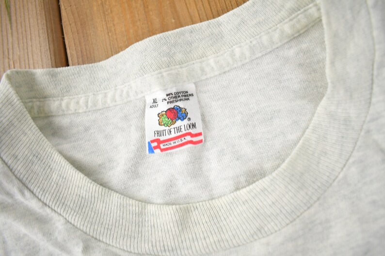 May include: A grey Fruit of the Loom t-shirt tag with the text "90% Cotton 10% Other Fibres Preshrunk" and "Fruit of the Loom" with a logo of fruit. The tag also says "XL Adult" and "Made in U.S.A."