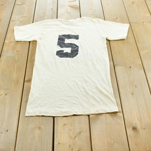May include: A short-sleeved, off-white t-shirt featuring a large, bold, black number 5 printed centrally on the chest. The garment has a crew neckline and a standard fit, displaying a simple design with a prominent numerical graphic.