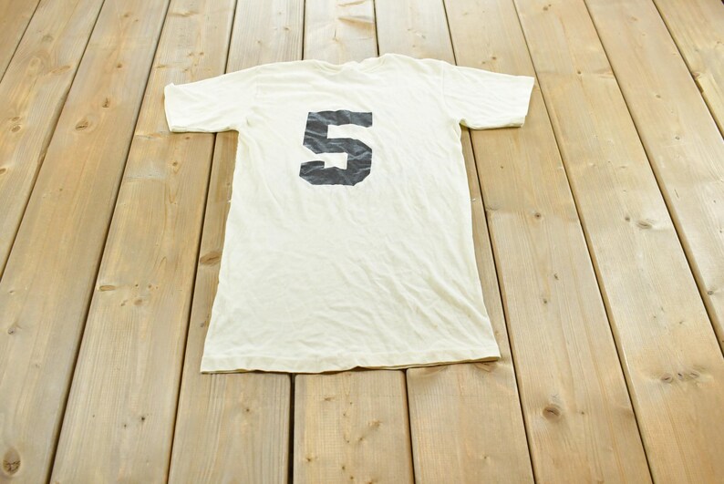 May include: A short-sleeved, off-white t-shirt featuring a large, bold, black number 5 printed centrally on the chest. The garment has a crew neckline and a standard fit, displaying a simple design with a prominent numerical graphic.