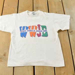 May include: White t-shirt featuring the phrase "WHAT WOULD JESUS DO?" in a colourful, block letter design. The tee has a crew neck and is made from a soft fabric. The text is in blue, orange, purple, and green.