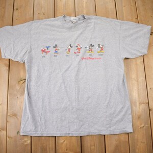 Puede incluir: Camiseta gris con un gráfico de Mickey Mouse en diferentes poses a lo largo de los años. El texto "Walt Disney World" está impreso debajo del gráfico.