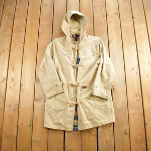 May include: A beige hooded  duffle coat with wooden toggles and a brown leather patch. The coat is laid flat on a wooden surface.