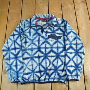 May include: Blue and white fleece pullover with a geometric pattern. The long-sleeved shirt has a button-down collar, a small chest pocket, and a navy blue trim. The Patagonia brand logo is visible on the chest pocket.