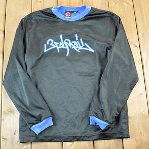 Puede incluir: Camiseta negra de manga larga con detalles azules en el cuello y los puños. La camiseta presenta la palabra "Bada$$" en una fuente estilizada de color azul claro en el pecho. La camiseta es de material ligero.