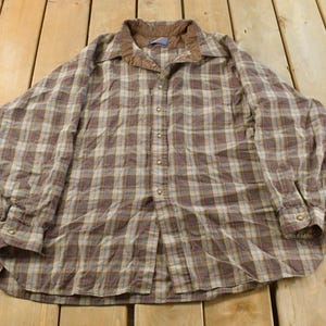 Puede incluir: Una camisa abotonada a cuadros marrones y beige con cuello de pana. La camisa de manga larga tiene un dise&ntilde;o cl&aacute;sico y est&aacute; hecha de un material suave y c&oacute;modo. La camisa est&aacute; tendida sobre una superficie de madera.