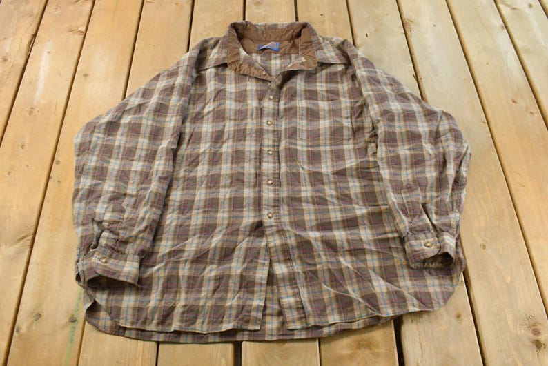 Puede incluir: Una camisa abotonada a cuadros marrones y beige con cuello de pana. La camisa de manga larga tiene un dise&ntilde;o cl&aacute;sico y est&aacute; hecha de un material suave y c&oacute;modo. La camisa est&aacute; tendida sobre una superficie de madera.