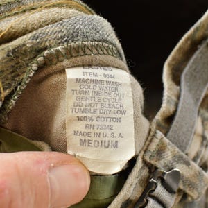 May include: Close-up of a camouflage-patterned garment with a tag. The tag reads "LADIES" and includes washing instructions: machine wash cold, tumble dry low. The garment is made of 100% cotton and is a medium size.