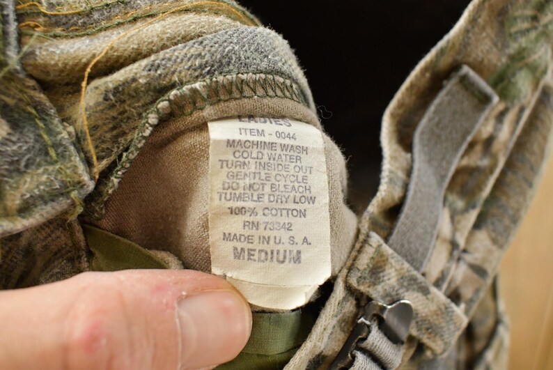 May include: Close-up of a camouflage-patterned garment with a tag. The tag reads "LADIES" and includes washing instructions: machine wash cold, tumble dry low. The garment is made of 100% cotton and is a medium size.