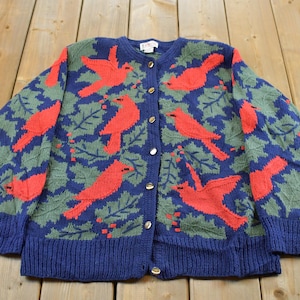 Puede incluir: Una chaqueta de punto azul marino con un patrón repetitivo de pájaros cardenales de color rojo brillante y hojas de acebo verdes. La prenda de manga larga tiene cuello redondo, cierre frontal con botones metálicos y puños y dobladillo acanalados.
