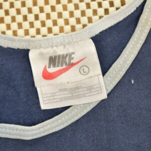 May include: A navy blue tank top with a white trim around the neckline. The tag on the shirt says "NIKE" in white letters with a red swoosh logo and "MADE IN U.S.A." in black text. The tag also has a size "L" in a white circle.